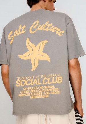 Muškarac nosi sivu majicu s žutim tekstom i grafikom morske zvijezde na kojoj piše "Salt Culture Social Club Sundays at the Beach, No Rules, Good Vibes."