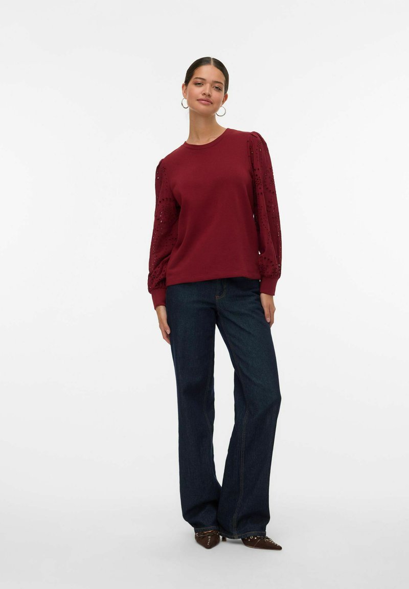 Sudadera burdeos con mangas abullonadas de textura similar a encaje, combinada con pantalones de mezclilla oscuros de pierna ancha y zapatos marrones de punta.