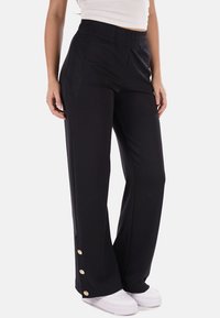 Pantaloni neri a gamba larga realizzati in tessuto morbido, con elastico in vita e bottoni dorati decorativi all'orlo. Abbinati a scarpe bianche.
