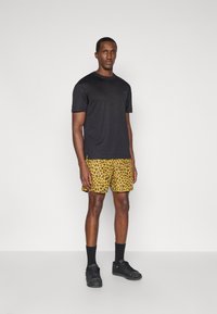 T-shirt preto de mangas curtas, calções amarelos com estampado de leopardo, meias pretas e sapatilhas. O conjunto é casual, apresentando cores e padrões atrevidos.
