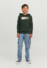 Felpa verde con una grafica bianca e arancione, tasca a marsupio, abbinata a jeans cargo blu chiaro con tasche laterali e sneakers.
