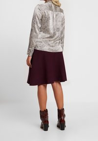 Blusa con motivo argento a maniche lunghe, abbinata a una gonna A-line color maroon. Modella indossa stivali alla caviglia marroni con tacco a blocco.