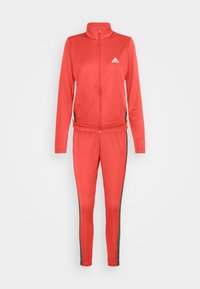 Survêtement rouge Adidas avec veste zippée intégrale et pantalon assorti comportant des bandes latérales noires, présenté sur un fond uni.