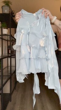 Šviesiai mėlyna satino blusa su sluoksniuotomis raukšlėmis, ilguis kaklo raiščiais ir šiek tiek blizgiu paviršiumi. Turi v formos iškirptę ir laisvas rankoves.