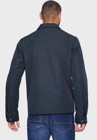 Mand iført en mørkeblå jakke og blå jeans, set bagfra mod en ensfarvet lys baggrund.