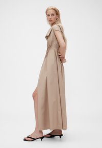 Beige klänning till anklarna med korta ärmar, krage och sidospalt. Sidenmjuk tyg med en slank silhuett, ihop med svarta sandaler.