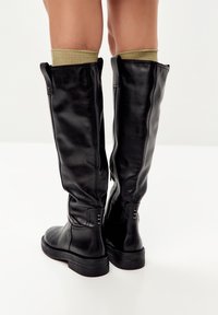 Botas de cuero negras hasta la rodilla con acabado mate, caña texturizada y suela de goma gris, que cuentan con cremalleras y acentos de calcetines de punto verde.