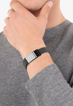 Pulsera de malla metálica en negro con un cierres de plata pulida que presenta un patrón de cuadrícula. Lleva en una muñeca, combinada con un suéter gris.