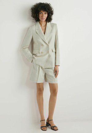 Kobieta z kręconymi włosami, ubrana w jasno beżową marynarkę dwurzędową i dopasowane krótkie spodenki sięgające kolan, stojąca na tle gładkiej białej ściany.