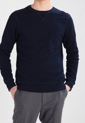 Man wearing a plain dark navy long-sleeve crew neck shirt with grey pants, hands casually in pockets, standing against a light background.