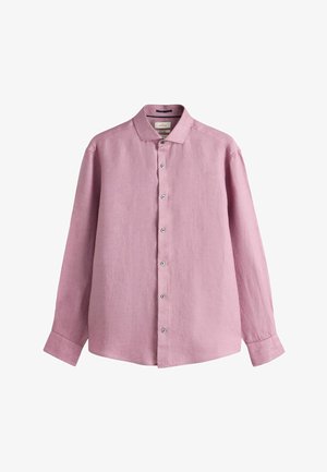 Stredoškolák černoch so sivou bradou, oblečený v mauve košeli na gombíky, pozerá priamo do kamery na jednofarebnom pozadí.