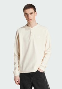 Camisa henley de manga larga color crema con tejido texturizado, dos botones en el cuello y un pequeño logo blanco en el pecho. Pantalones negros.