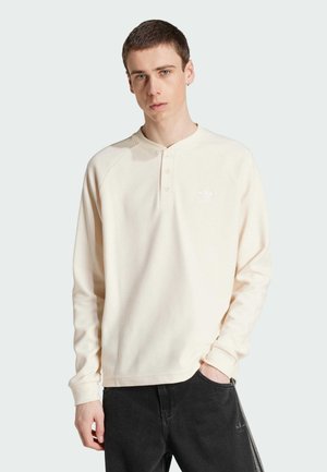 Krem bijela dugorukavna henley majica s teksturiranim materijalom, dva gumba na vratu i mali bijeli logo na prsima. Crne hlače.