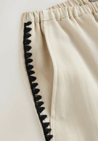 Pantaloni in tessuto beige con vita elasticizzata e rifinitura ricamata triangolare nera lungo la cucitura laterale vicino alla tasca.
