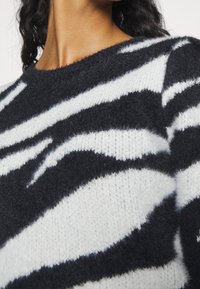 Pull rayé noir et blanc en maille douce, présentant un col rond et une texture duveteuse avec des motifs ondulés audacieux.