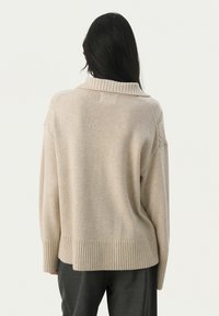 Beige knit sweater with a high ribbed collar, dropped shoulders, and ribbed cuffs. Soft texture, relaxed fit, and plain design.