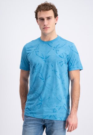 Jonge man met krullend haar die een blauw T-shirt met patroon en korte mouwen draagt en een lichtblauwe spijkerbroek, tegen een effen lichte achtergrond.