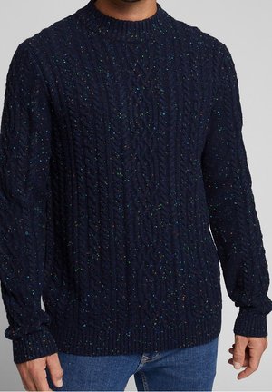 Marineblauer Strickpullover mit Zopfmuster und mehrfarbigen Sprenkeln, getragen zu blauen Jeans, sichtbar vom Hals bis zu den Hüften.