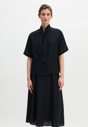 Vrouw draagt een zwarte blouse met korte mouwen en een geknoopt neksjaaltje, met een lange zwarte rok, staand tegen een effen witte achtergrond.