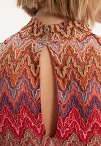 Spitzenoberteil mit einem Zickzackmuster in warmen Farben, darunter Rot, Orange und Blau. Verfügt über einen hohen Ausschnitt und einen Schlüsselloch-Rücken mit Knöpfen.