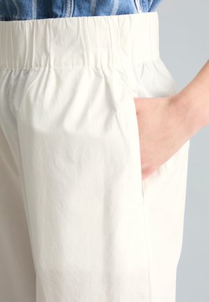 Main dans la poche d’un pantalon blanc taille haute avec taille élastique, porté avec une veste en jean rentrée.