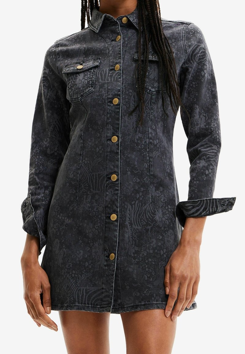 Robe cintrée en denim foncé avec un imprimé animal discret, fermeture à boutons sur le devant, poches poitrine, manches longues retroussées aux poignets, portée par une personne aux cheveux tressés.