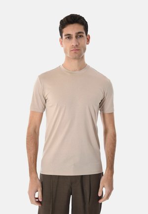 Uomo che indossa una t-shirt beige a maniche corte con scollo a girocollo e pantaloni marrone scuro, in piedi contro uno sfondo bianco semplice.