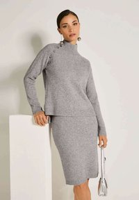 Frau, die einen grauen Strickpullover mit Knöpfen an der Schulter und einen passenden Strickrock trägt, während sie eine metallische Handtasche hält.