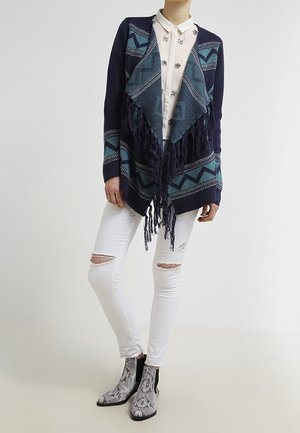Frau trägt eine marineblaue und türkis gemusterte Fransen-Cardigan, ein weißes, verziertes Hemd mit Knöpfen, zerrissene weiße Skinny-Jeans und Stiefeletten mit Schlangenprint.