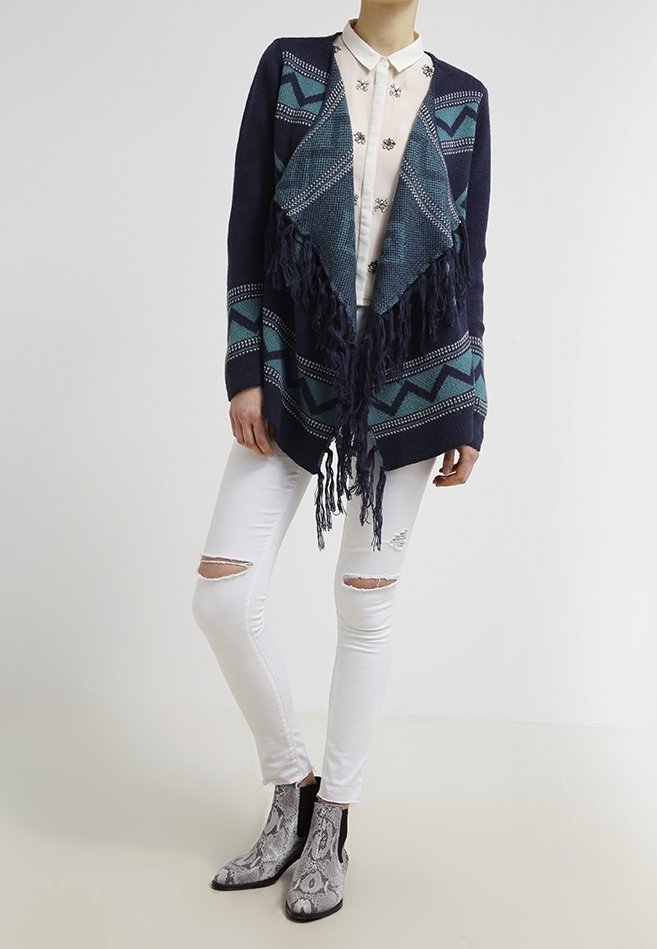 Femme portant un cardigan à franges bleu marine et bleu sarcelle à motifs, une chemise blanche boutonnée ornée, un jean skinny blanc déchiré et des bottines à imprimé serpent.