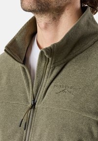 Päivänvihreä fleece-takki, jossa on korkea kaulus, vetoketjullinen etuosa ja brodeerattu logo. Teksturoitu kangas ja kontrastivärinen sisäkauluksen yksityiskohta.