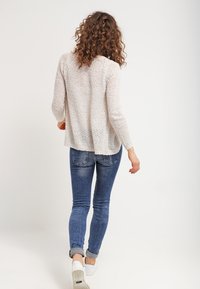 Ljusbeige texturerad stickad tröja med långa ärmar, bärs med blå skinny jeans och vita sneakers, sedd bakifrån.