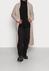 Cardigan tricoté beige à l'avant ouvert, surface texturée, assorti d'un col roulé noir et d'un pantalon, avec des chaussures épaisses noires.