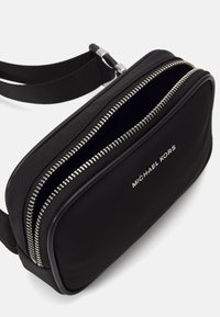 Sac bandoulière en nylon noir avec une fermeture éclair argentée, un accent logo et une texture lisse. Forme rectangulaire compacte avec une sangle ajustable.