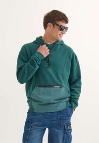 Sudadera verde con un acabado texturizado, bolsillo en contraste más claro, capucha con cordón y acento del logo en negro. Combinada con jeans cargo azules.