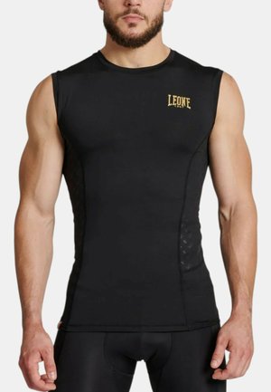 Uomo muscoloso che indossa una maglia atletica nera aderente senza maniche con logo "LEONE 1947" sul petto, in piedi davanti a uno sfondo bianco.