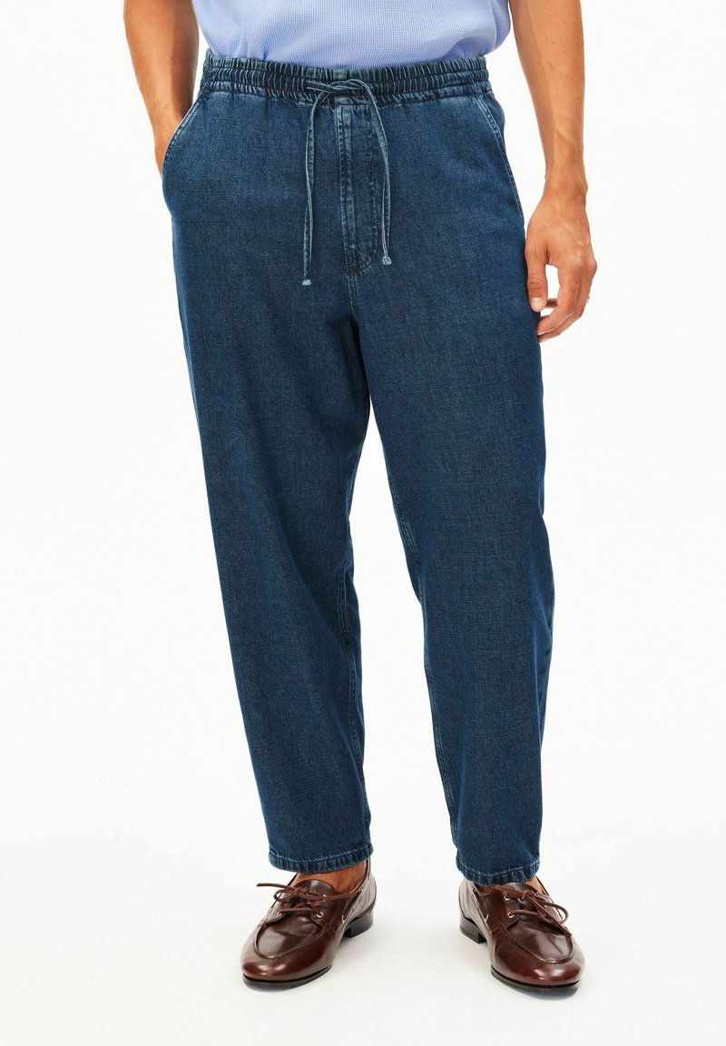 Mann trägt dunkelblaue Denim-Hose mit elastischem Bund und Kordelzug, kombiniert mit braunen Leder-Schnürschuhen und hellblauem Hemd.