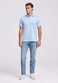 Lichtblauwe polo met een kraag en drie knopen, gedragen met lichtdenim jeans en witte sneakers. De stof lijkt soepel en iets getailleerd.