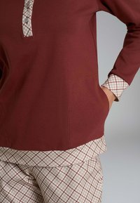 Top color amaranto con scollo abbottonato, caratterizzato da un bordo a contrasto a quadri. La parte inferiore è in tessuto beige con motivo a scacchiera.