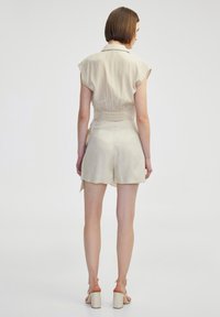 Beige, korte jumpsuit met korte mouwen en een cinched taille. Zachte stof, detail met strik op de rug en een relaxte pasvorm. Gecombineerd met chunky sandalen met een hak.