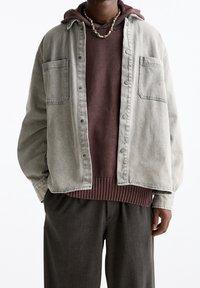 Szara kurtka dżinsowa z zatrzaskami, dwoma kieszeniami na piersi, nałożona na brązowy hoodie, zestawiona z ciemnymi, teksturowanymi spodniami. Naszyjniki dodają detali.