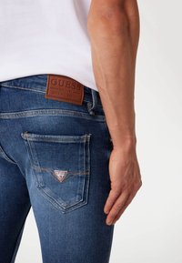 Blauwe denim jeans met een bruine leren patch met merknaam, driehoek logo op de achterzak en een getailleerd ontwerp met zichtbare stiksels.