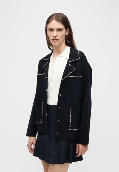 Νεαρή γυναίκα με μαύρο τζάκετ με καρφιά πάνω από λευκό πουκάμισο με κουμπιά και μαύρη πλισέ φούστα, που στέκεται μπροστά σε απλό φόντο.