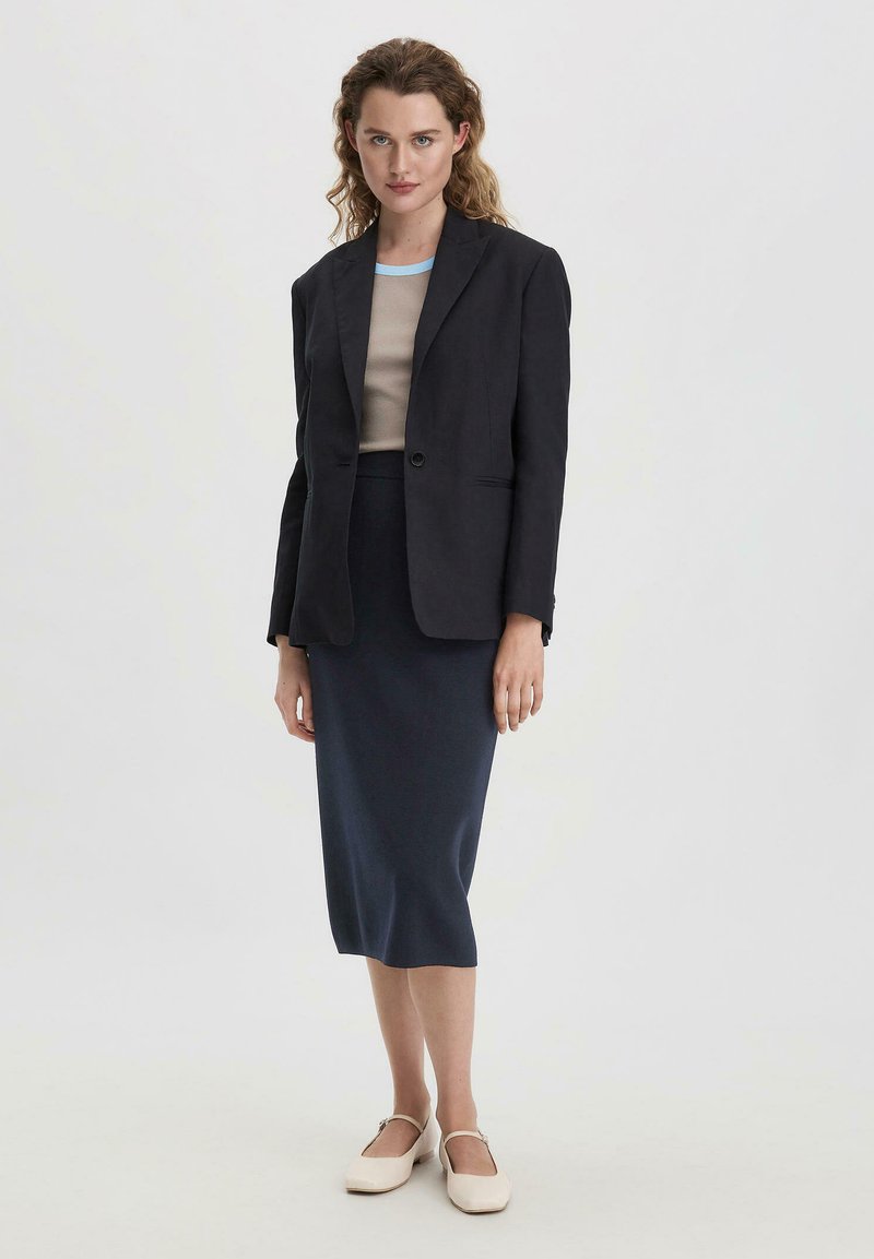 Marineblaue Blazer mit taillierter Passform, beiges Oberteil mit blauen Akzenten und knielanger dunkler Rock. Ergänzt durch hellfarbige Slipper.