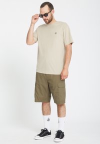 Beżowa koszulka z krótkim rękawem z małym logo, zestawiona z oliwkowymi szortami typu cargo. Całość uzupełniają czarne sneakersy i białe skarpetki.