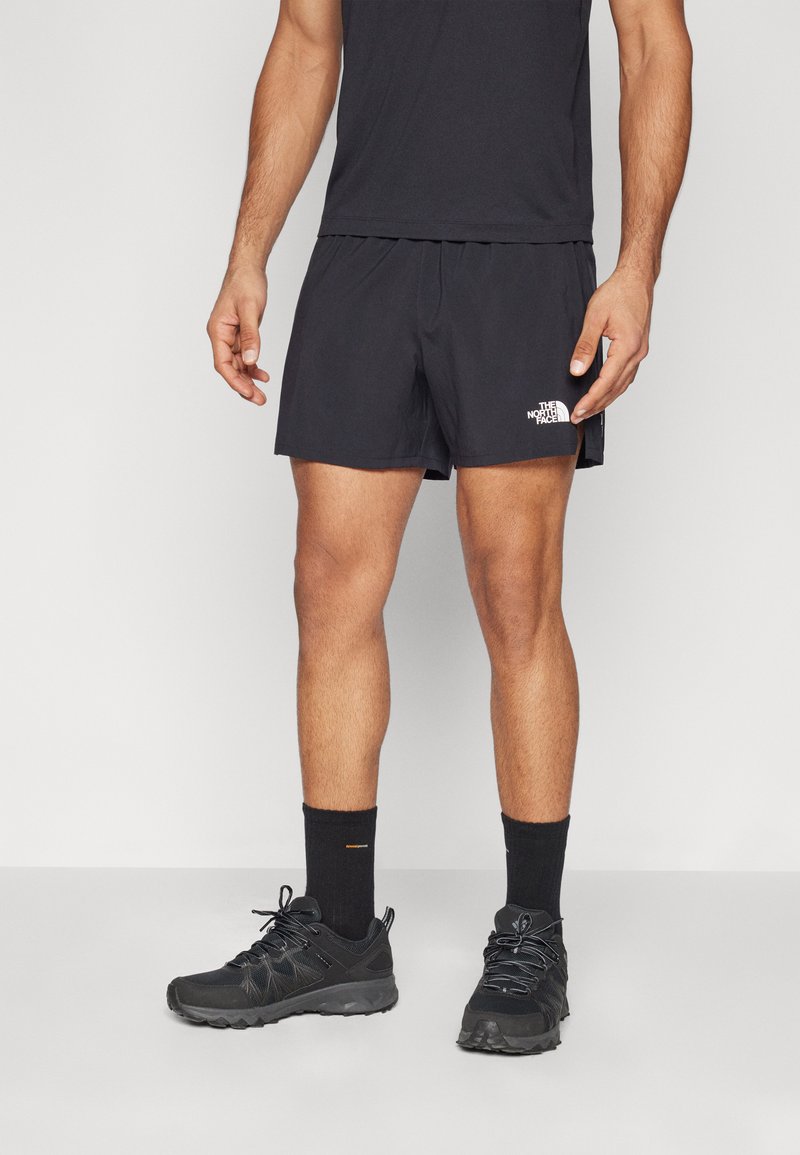 Svarte treningshorts med elastisk midjebånd, med en hvit logo på venstre side. Kombinert med svarte sko og sokker.