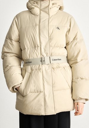 Giacca invernale imbottita beige con cappuccio, cintura elastica Calvin Klein, tasche e logo CK sul petto, indossata sopra pantaloni neri.