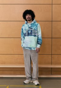 Hanorac cu glugă cu un model albastru și mentă, prevăzut cu un buzunar frontal și șnur; combinat cu pantaloni din velur bej deschis și adidași.