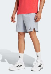 Los pantalones cortos deportivos de color gris claro cuentan con un logo de tres franjas en negro, un corte relajado y ventilaciones laterales para mayor movilidad, combinados con zapatos y calcetines blancos.