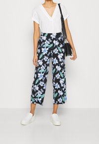 Pantalons à imprimé floral sur fond noir, avec des fleurs bleues et vertes. Associés à un haut blanc à manches courtes et des chaussures blanches à enfiler.